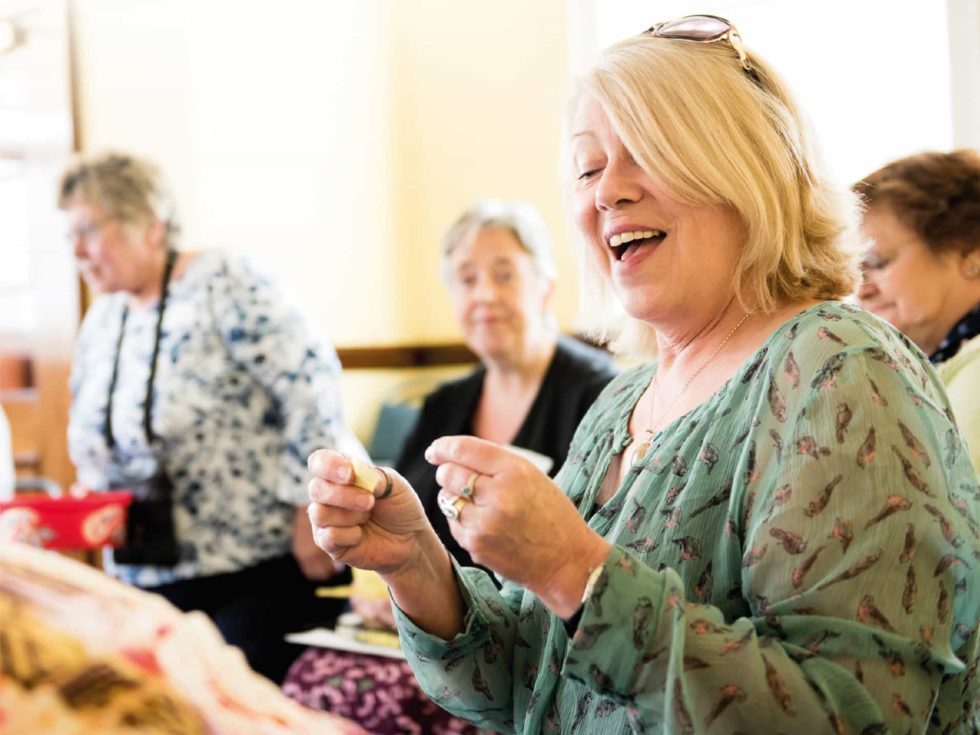 British Lung Foundation - Helping Britain breathe better. Lady wearing a green blouse holding a peice of paper gently laughing with three other ladies in the background.