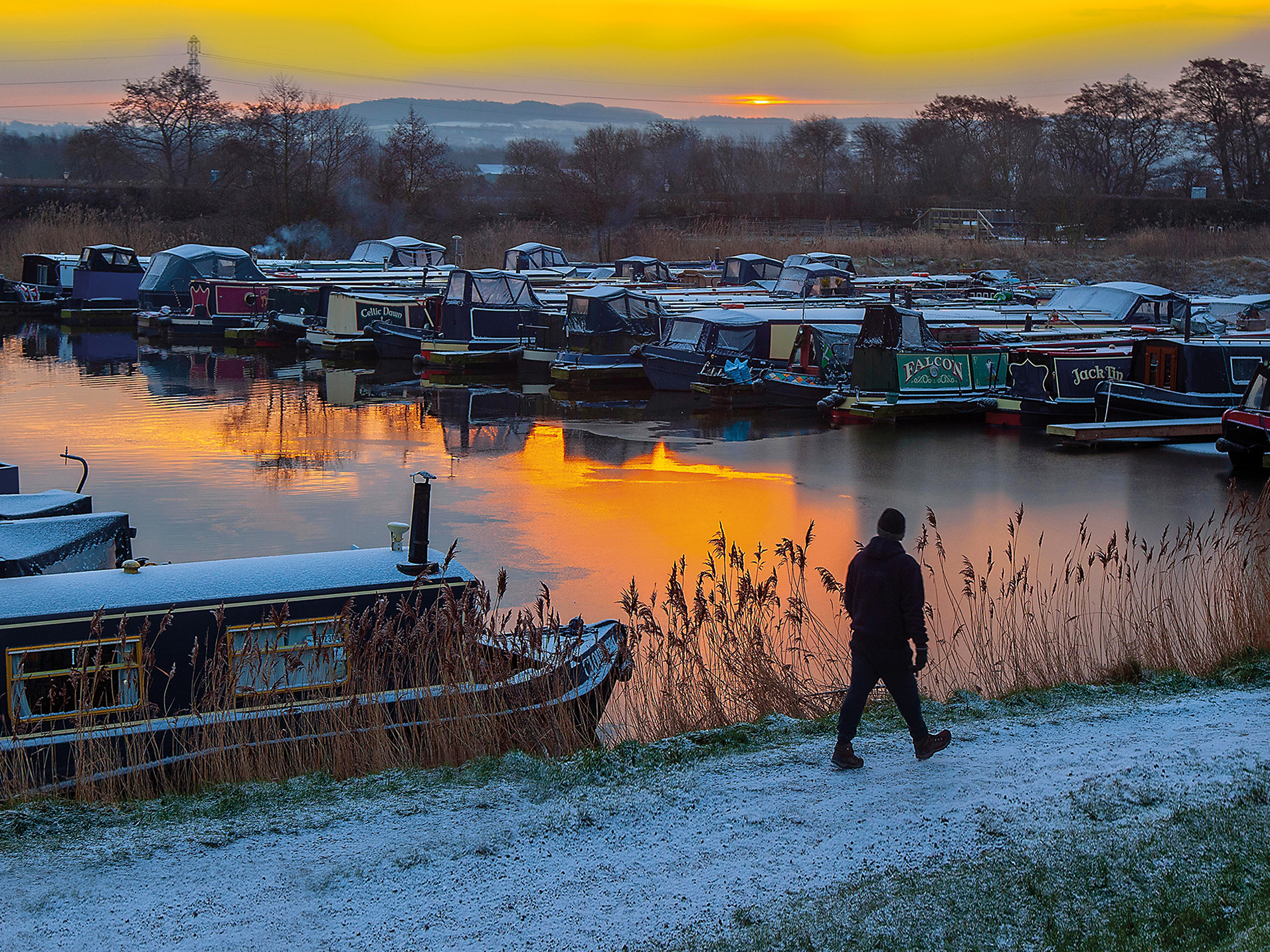 Canal & River Trust - Friends Waterfront magazine and emails. Man walking along a riverbank while on a pisturesque winter morning while looking at a group of moored boats.